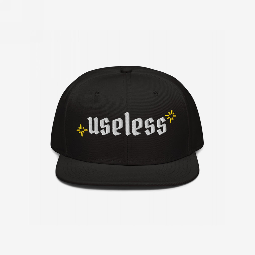 The Useless Hat features a flat-bill snapback design, with useless embroidered in white Old English font, flanked by yellow starbursts, embodying a relaxed, rebellious spirit.
