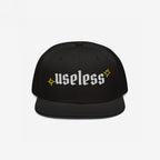 The Useless Hat features a flat-bill snapback design, with useless embroidered in white Old English font, flanked by yellow starbursts, embodying a relaxed, rebellious spirit.