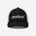 The Useless Hat, a two-tone trucker cap, features useless in bold Old English font on black fabric, flanked by yellow starbursts, embodying a chill, revolutionary vibe.