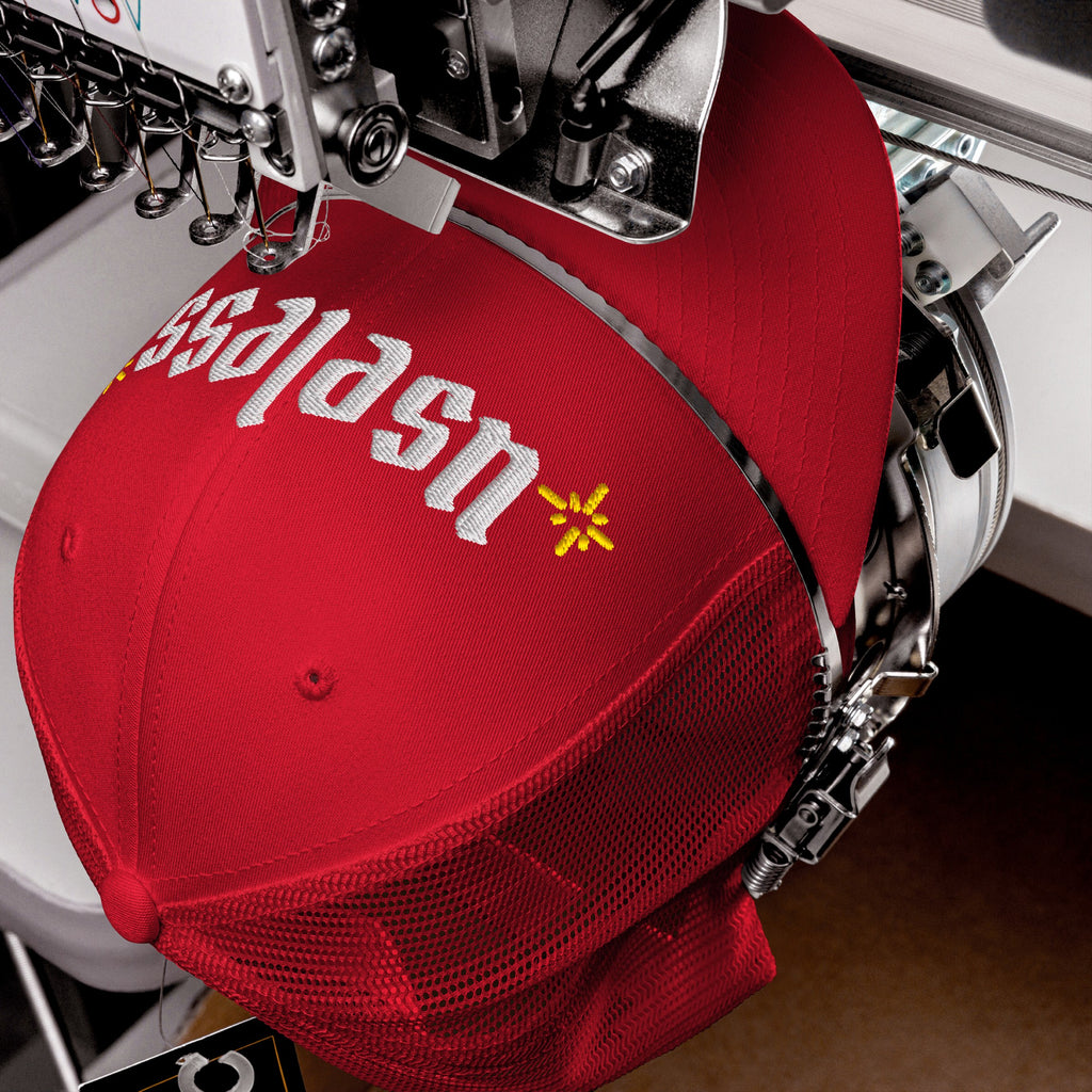 The Useless Hat, a red trucker cap, is in an embroidery machine, stitching sslash and a yellow star-burst, symbolizing rest and resistance against hustle culture.