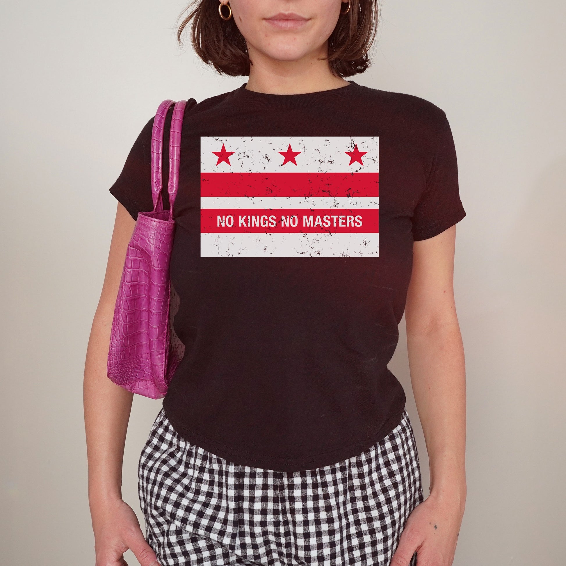 Person wearing the Washington DC No Kings Flag Women's Baby Tee with distressed red-and-white graphic, paired with black-and-white gingham trousers and a purple shoulder bag, embodying a bold anti-authority statement.