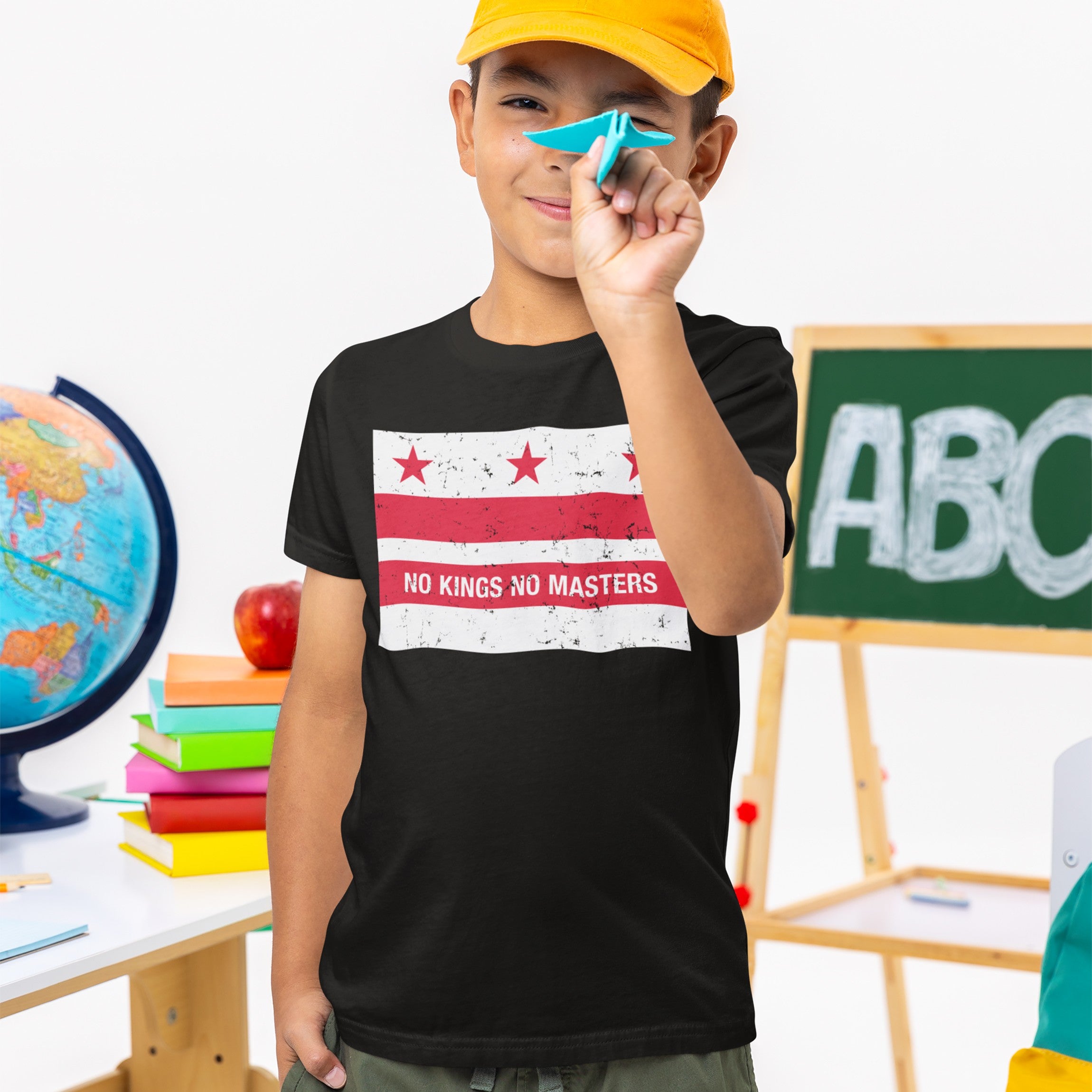 Boy in classroom holds a blue paper airplane while wearing Washington DC No Kings Flag Shirt, featuring red stars and stripes with the slogan “NO KINGS NO MASTERS.” Background includes educational items.