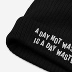 The Wasted Days Hat features a black rib-knit beanie with a folded cuff, embroidered with the phrase, “A DAY NOT WASTED IS A DAY WASTED,” embodying a bold, carefree philosophy.