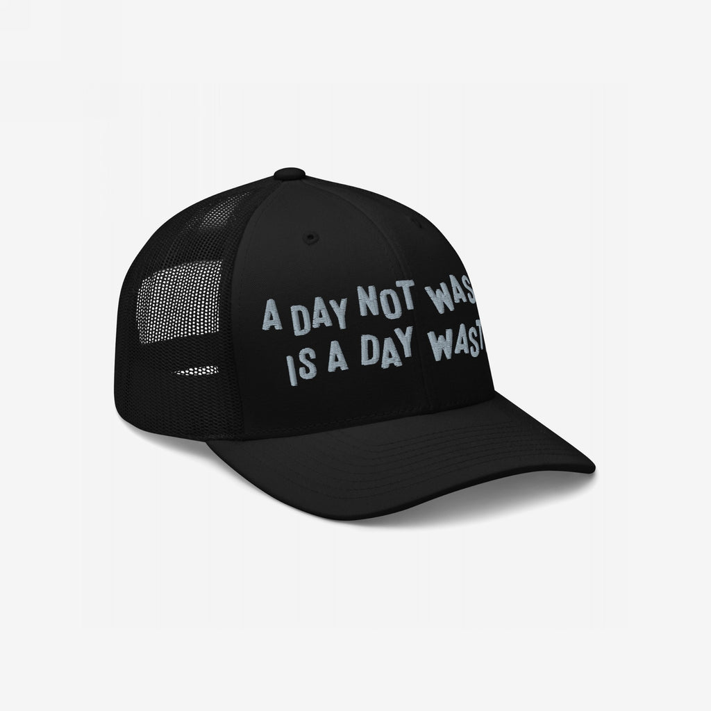 Black trucker cap features white embroidery: A Day Not Wasted Is A Day Wasted. Solid front with mesh back, embodying a bold, carefree lifestyle. Ideal for lively gatherings and spirited adventures.