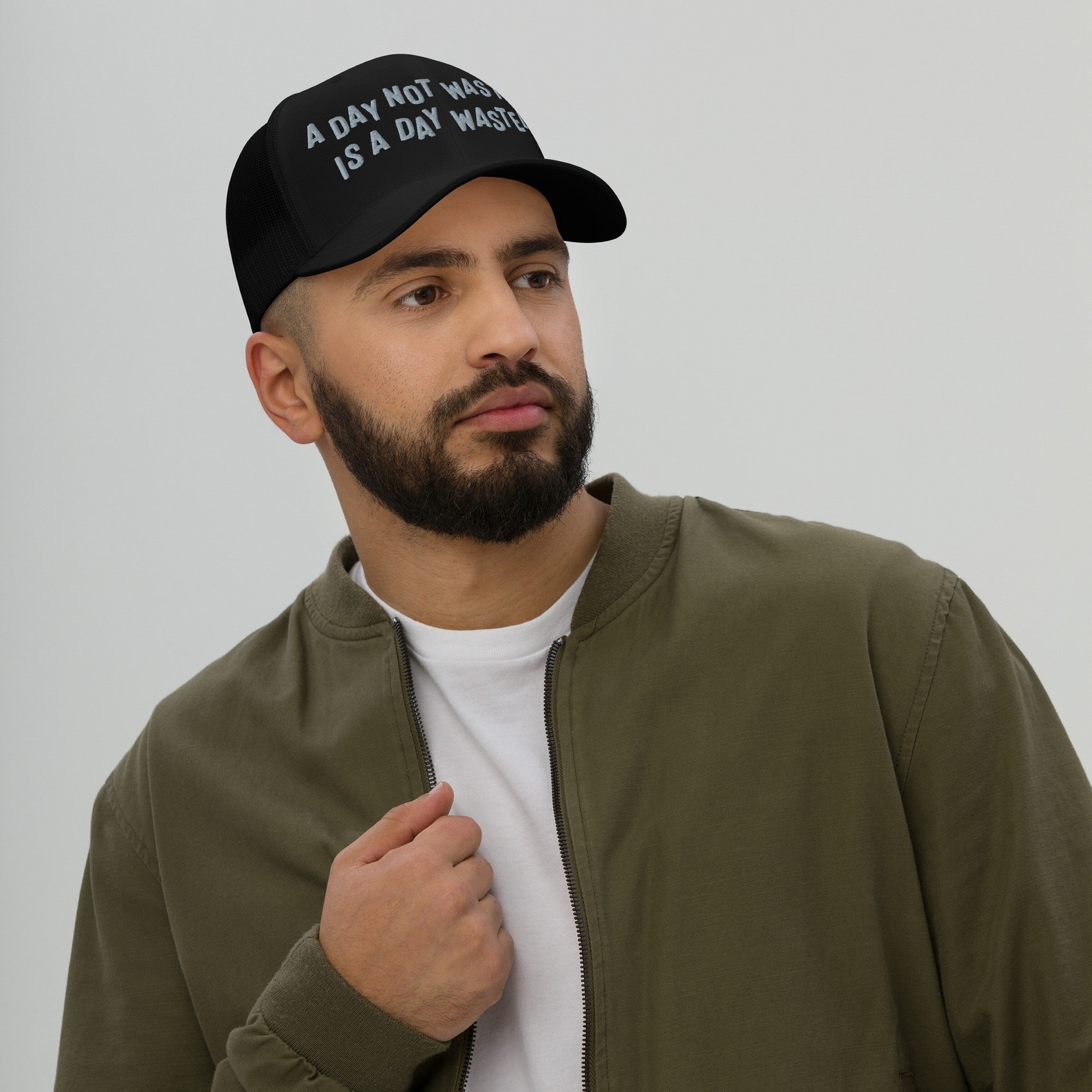 Close-up of a bearded man wearing the Wasted Days Hat, a black trucker-style cap featuring the bold slogan “A DAY NOT WASTED IS A DAY WASTED” in white, embodying carefree, adventurous spirit.