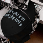 Wasted Days Hat being embroidered with “A DAY NOT WASTED” on a black cap by a machine, capturing the carefree and bold essence of living unapologetically.