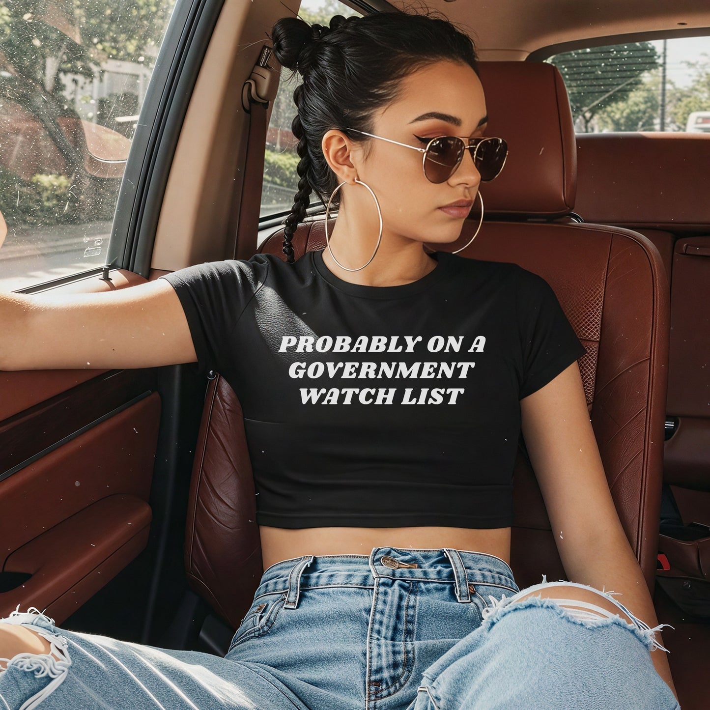 Young woman in aviator sunglasses and braided buns wears the Watch List Women's Baby Tee, a black cropped shirt with bold text, embodying a rebellious, activist spirit, paired with light-wash, ripped jeans.