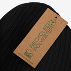 Close-up of the Watch List Hat, a black rib-knit beanie with a recycled polyester tag, designed for activism and visibility. Features a green-lettered tag from ATLANTIS, emphasizing sustainability and protest messaging.