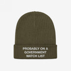 The Watch List Hat features an olive-green, ribbed knit design with a folded cuff, embroidered with “PROBABLY ON A GOVERNMENT WATCH LIST,” reflecting bold activism and individuality.