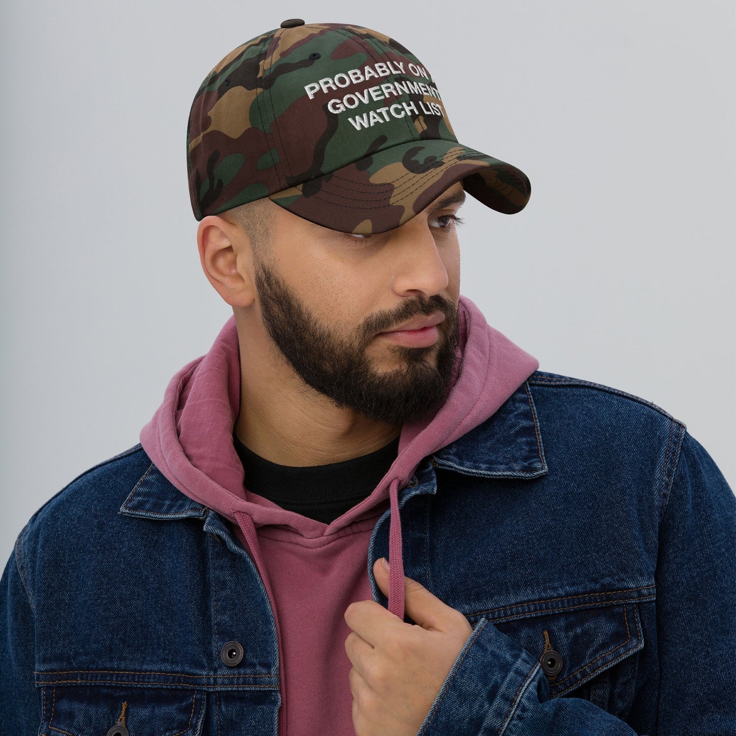 Bearded man wearing the Watch List Hat, a camouflage baseball cap with PROBABLY ON GOVERNMENT WATCH LIST slogan; paired with a hoodie and denim jacket, embodying bold protest statements.