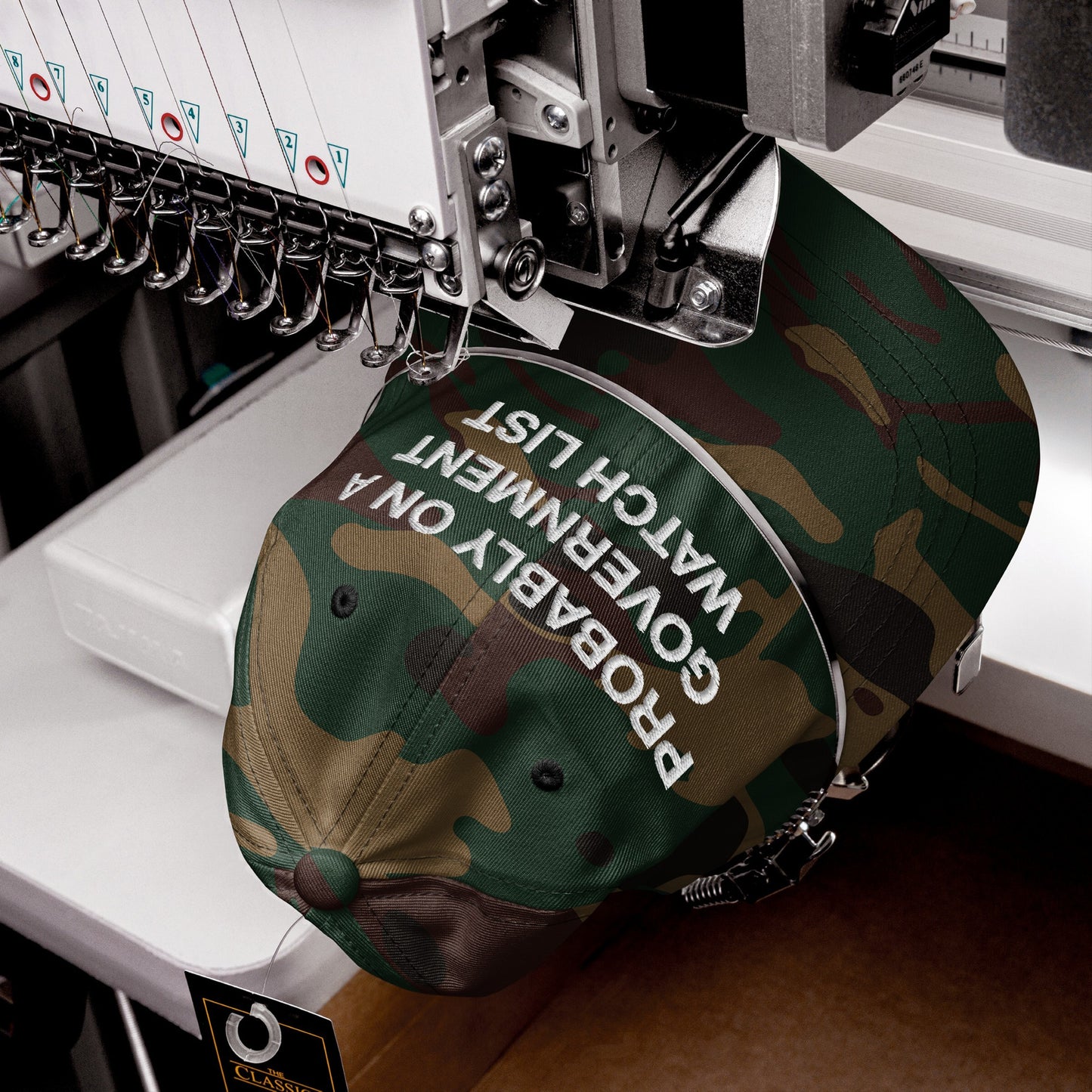 Close-up of the Watch List Hat, an industrial embroidery machine stitching “PROBABLY ON A GOVERNMENT WATCH LIST” onto a camouflage baseball cap, embodying bold defiance and civic responsibility.