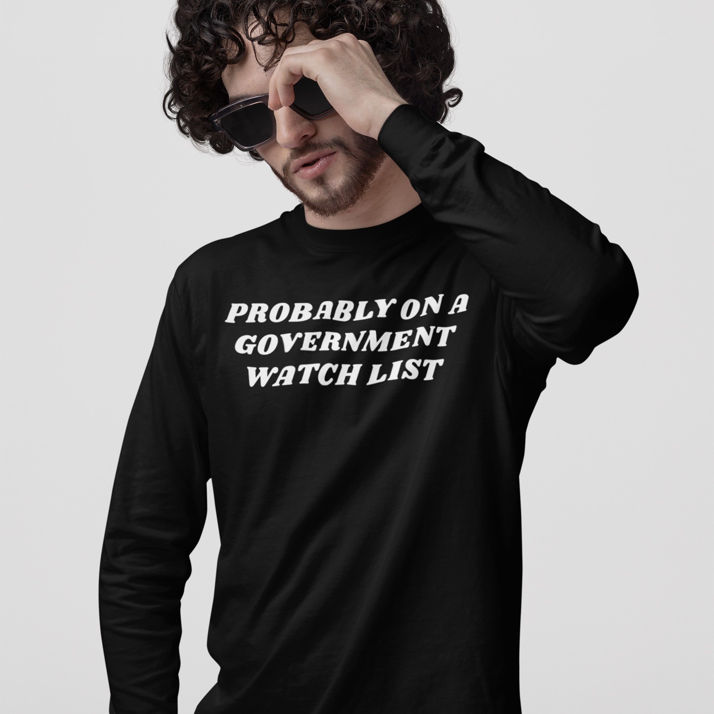 Young man with curly hair adjusts sunglasses, wearing a Watch List T-shirt featuring the bold slogan, “PROBABLY ON A GOVERNMENT WATCH LIST,” reflecting defiance and political activism.