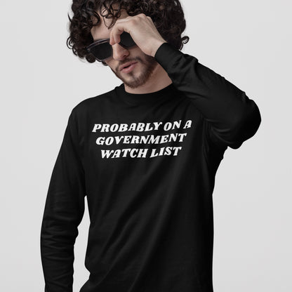 Young man with curly hair adjusts sunglasses, wearing a Watch List T-shirt featuring the bold slogan, “PROBABLY ON A GOVERNMENT WATCH LIST,” reflecting defiance and political activism.