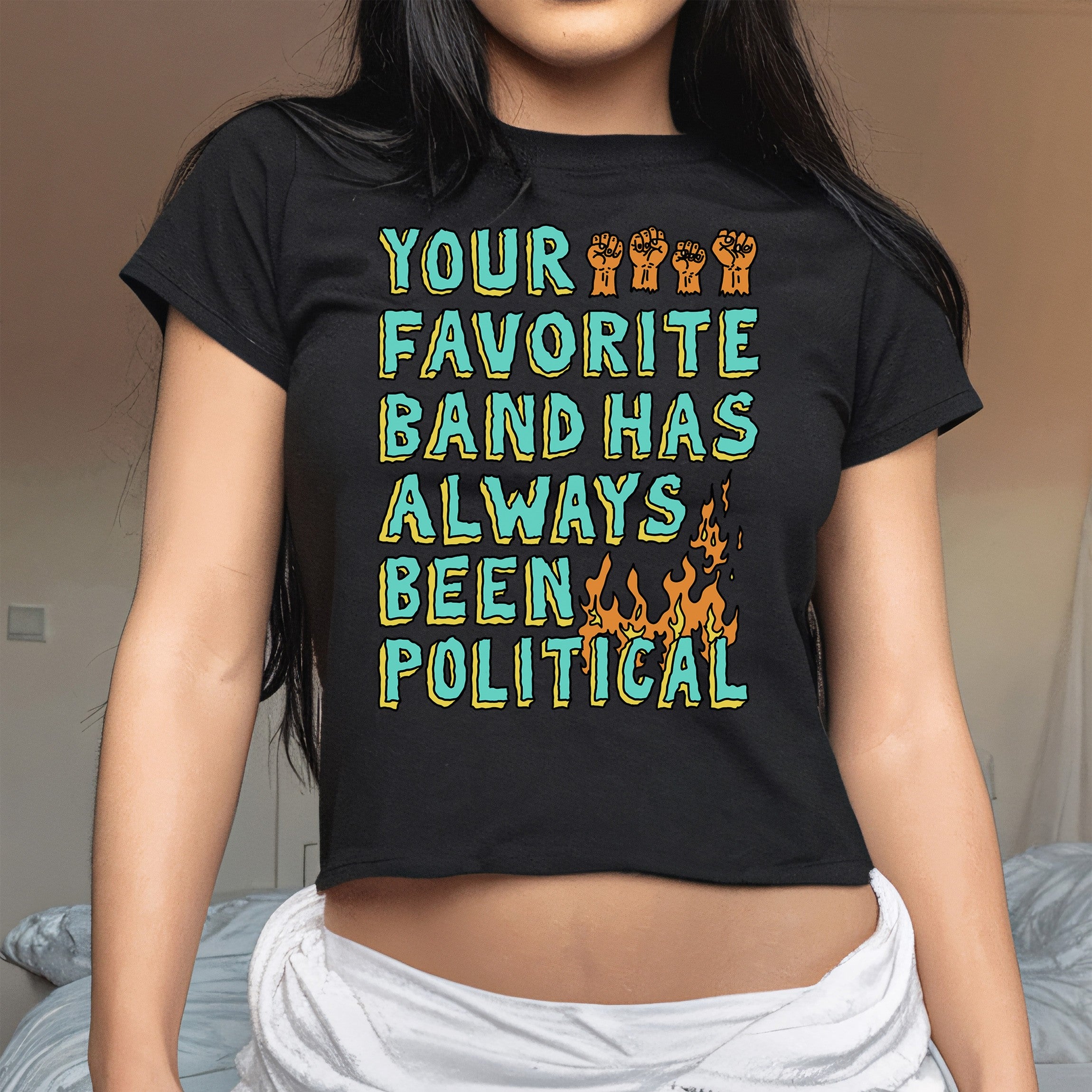 Your Favorite Band Has Always Been Political Women's Baby Tee featuring bold text, raised-fist illustrations, and stylized flames, worn casually indoors, embodying punk's inseparable link to politics.